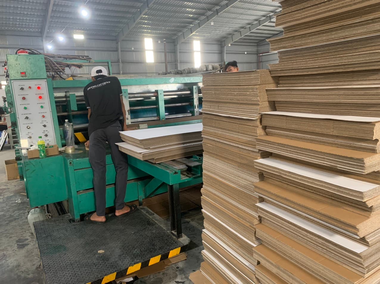 Staff operating the automatic horizontal printing machine beside stacks of carton sheets at the Phuong Cat factory